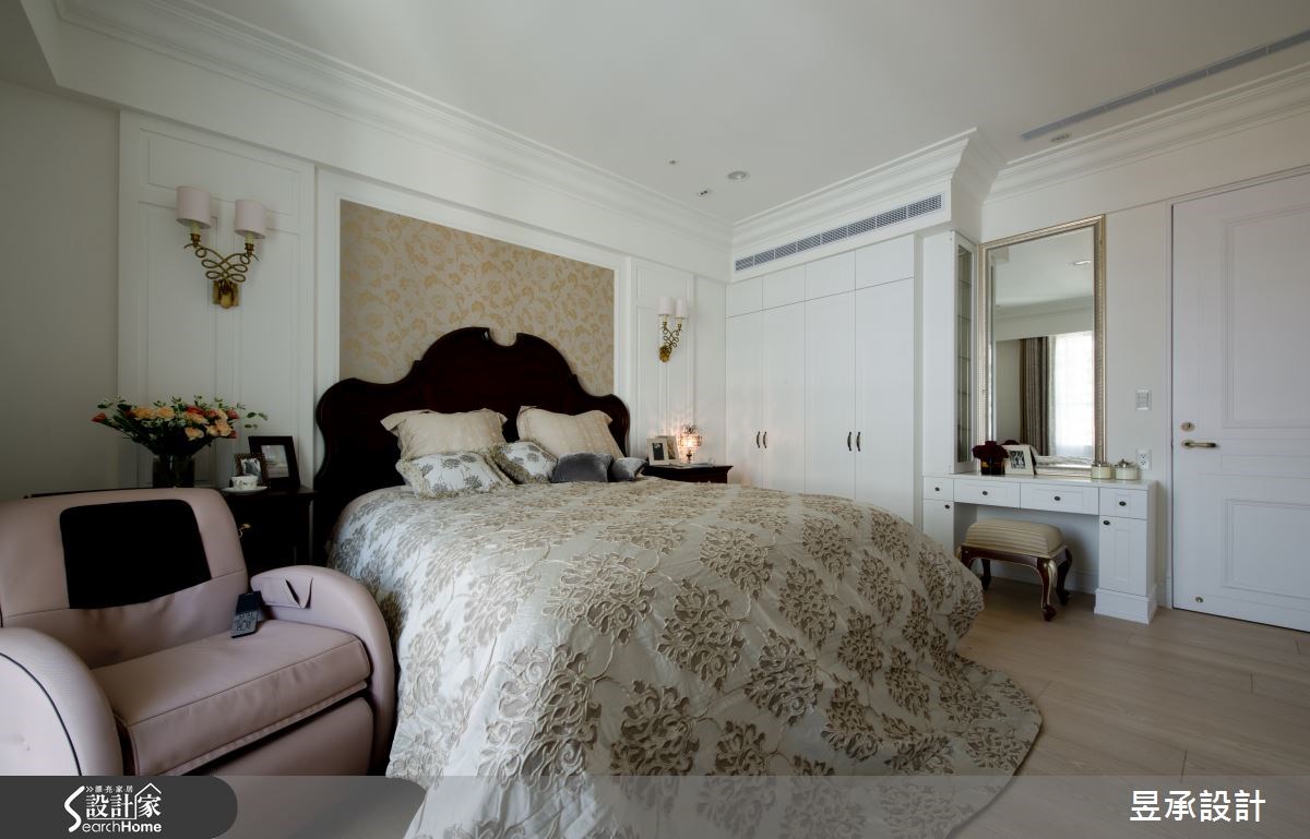 主臥室床頭以白色對稱設計的古典壁板加上金色的古典圖藤壁紙，點出了空間的優雅調性；化妝桌採側邊滑軌的收納設計，不但滿足屋主的生活需求，更貼心考量到古典畫框鏡的整體視覺美感。
